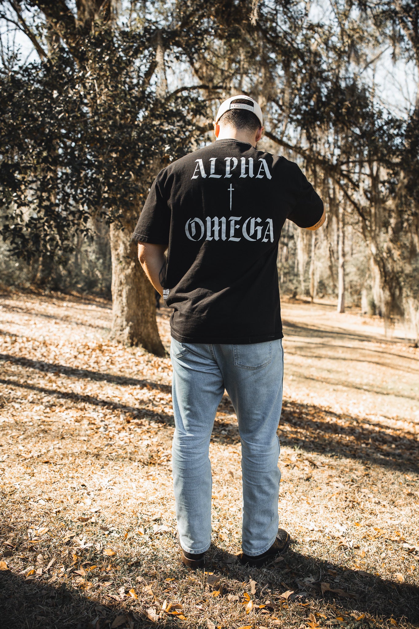 A man stands outdoors on dry grass facing away, wearing the 13th Alpha & Omega Premium Oversized Tee - Black with "Alpha + Omega" printed on the back, styled with light blue jeans, black shoes, and a beige cap among trees—Christian streetwear.