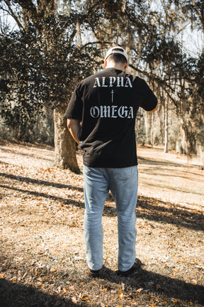 A man stands outdoors on dry grass facing away, wearing the 13th Alpha & Omega Premium Oversized Tee - Black with "Alpha + Omega" printed on the back, styled with light blue jeans, black shoes, and a beige cap among trees—Christian streetwear.