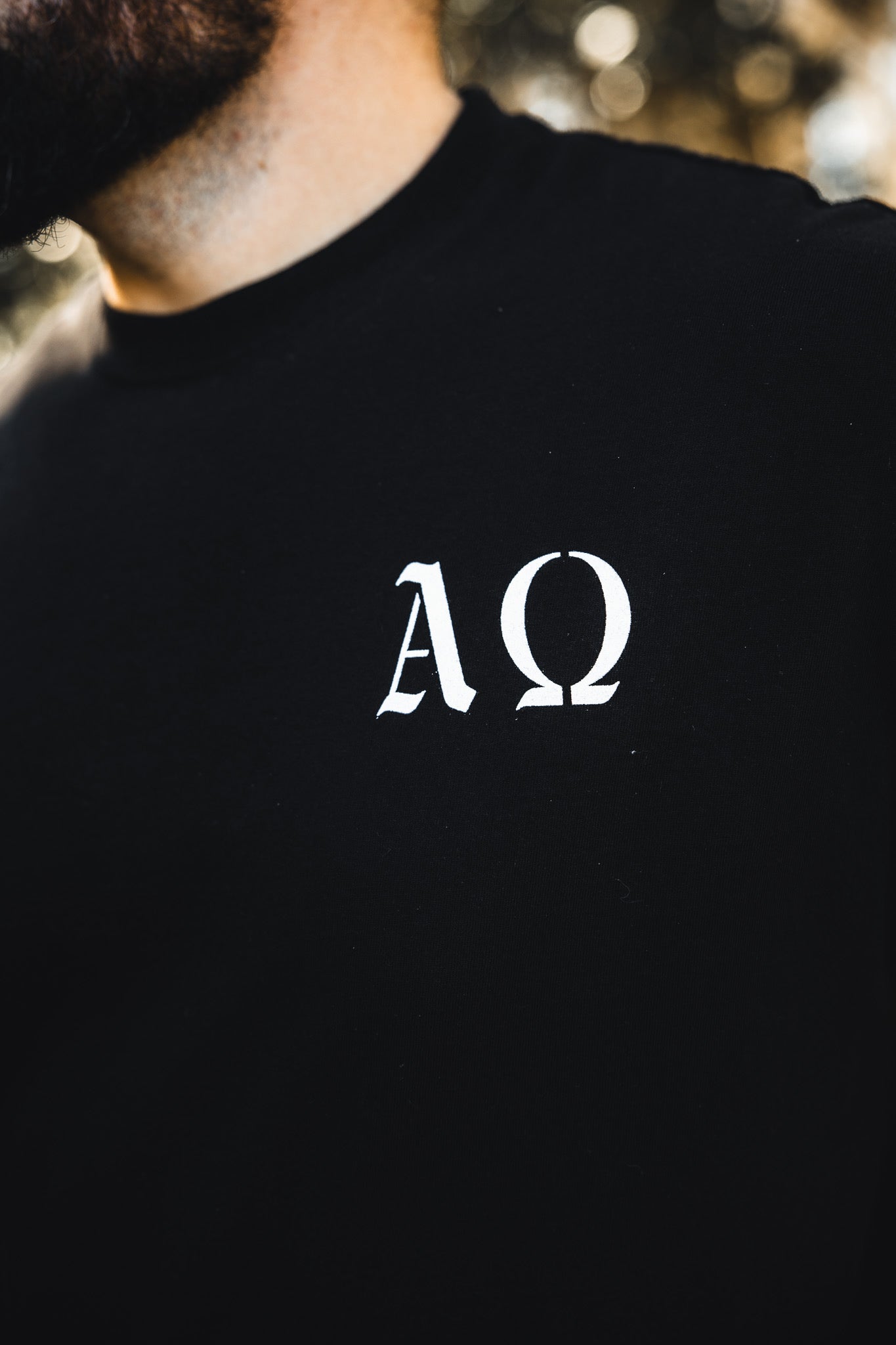 Close-up of a person wearing the 13th Alpha & Omega Premium Oversized Tee - Black, crafted from heavyweight cotton and featuring white Greek letters Α and Ω on the front. The face is hidden—classic Christian streetwear style.