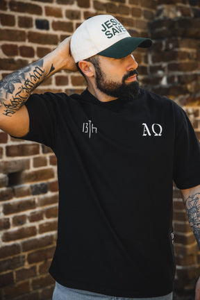 A bearded man in a white Jesus Saves cap and the 13th Alpha & Omega Premium Oversized Tee - Black poses before a brick wall, showcasing his tattooed arms—a striking example of bold Christian streetwear.