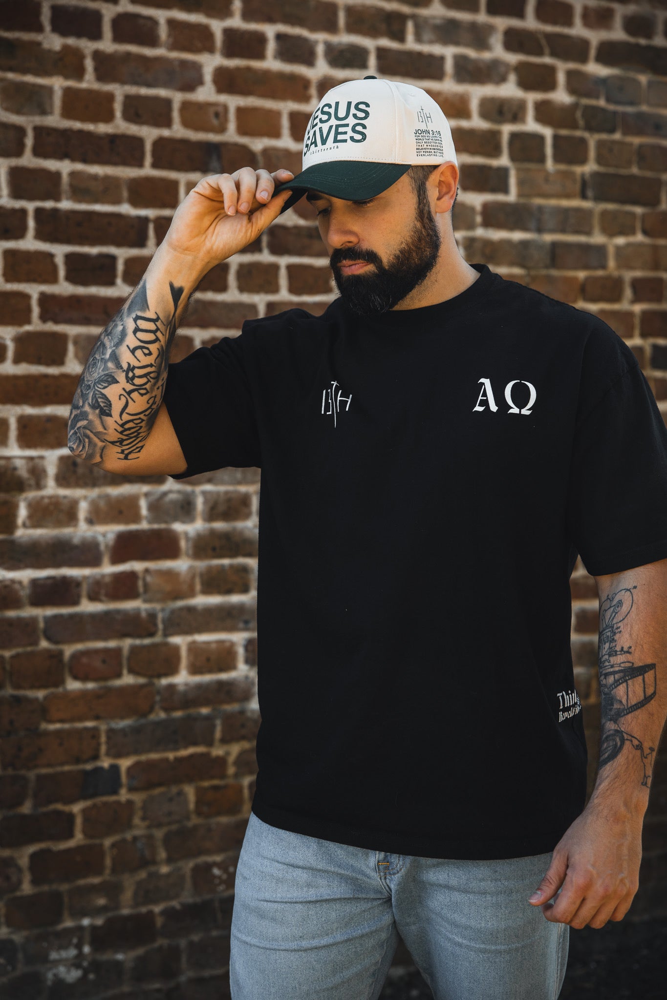 A bearded, tattooed man in a black T-shirt stands by a brick wall, tilting the brim of the 13th Jesus Saves Ivory Snapback featuring John 3:16 details.
