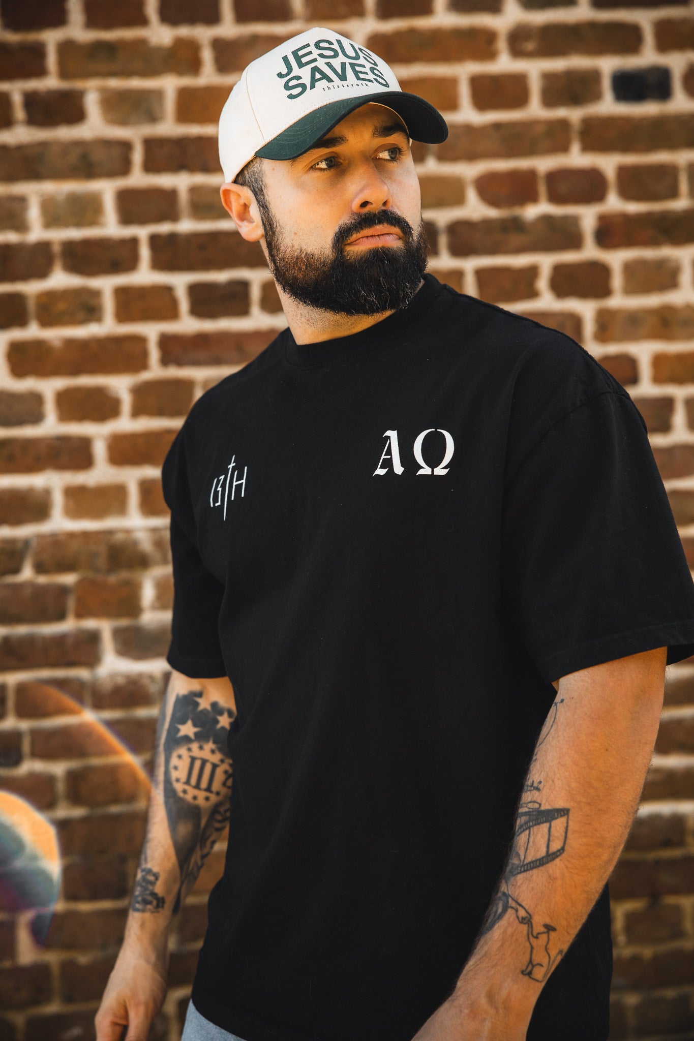A bearded, tattooed man stands before a brick wall, wearing a black tee with white symbols and the 13th Jesus Saves Ivory Snapback, featuring "JESUS SAVES" in green letters—a reference to John 3:16. He gazes off to the side.