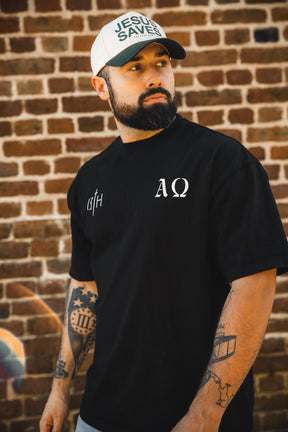 A bearded, tattooed man stands before a brick wall, wearing a black tee with white symbols and the 13th Jesus Saves Ivory Snapback, featuring "JESUS SAVES" in green letters—a reference to John 3:16. He gazes off to the side.