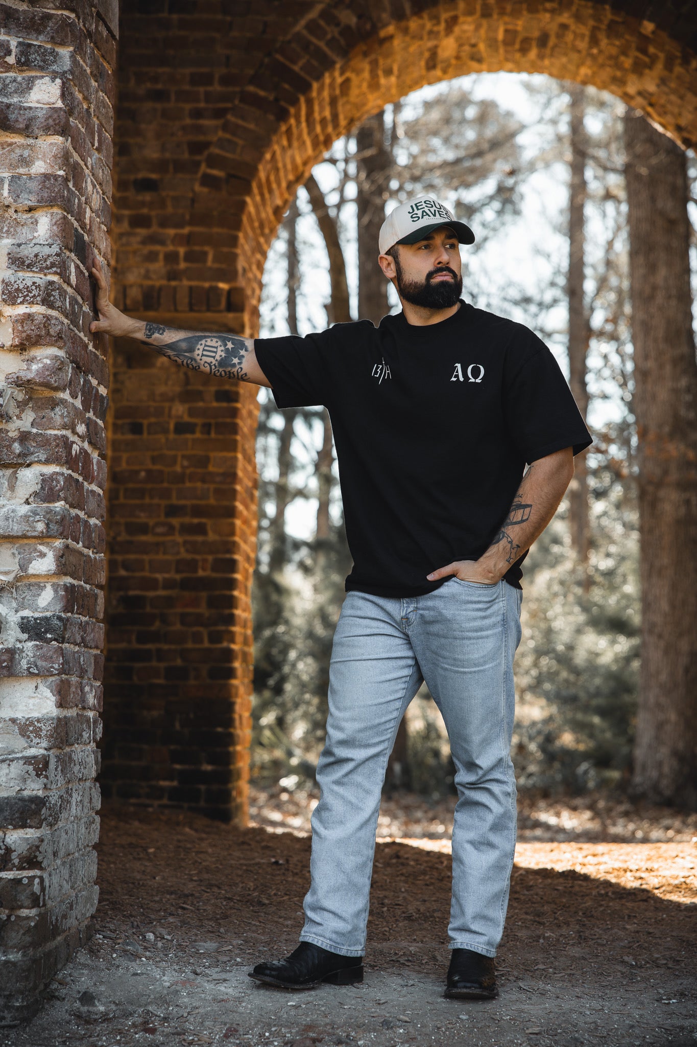A bearded man wearing the 13th Alpha & Omega Premium Oversized Tee in black, paired with light jeans and a cap, leans against a brick wall under an archway outdoors with trees in the background.