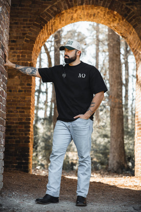A bearded man wearing the 13th Alpha & Omega Premium Oversized Tee in black with light jeans stands outdoors under a brick archway, sunlight filtering through trees and adding a relaxed vibe to his Christian streetwear look.