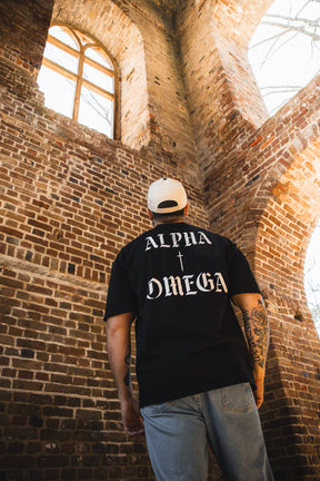 A person wearing the Alpha & Omega Premium Oversized Tee - Black by 13th and a white cap stands inside a brick building with arched windows, gazing upward as sunlight pours in.
