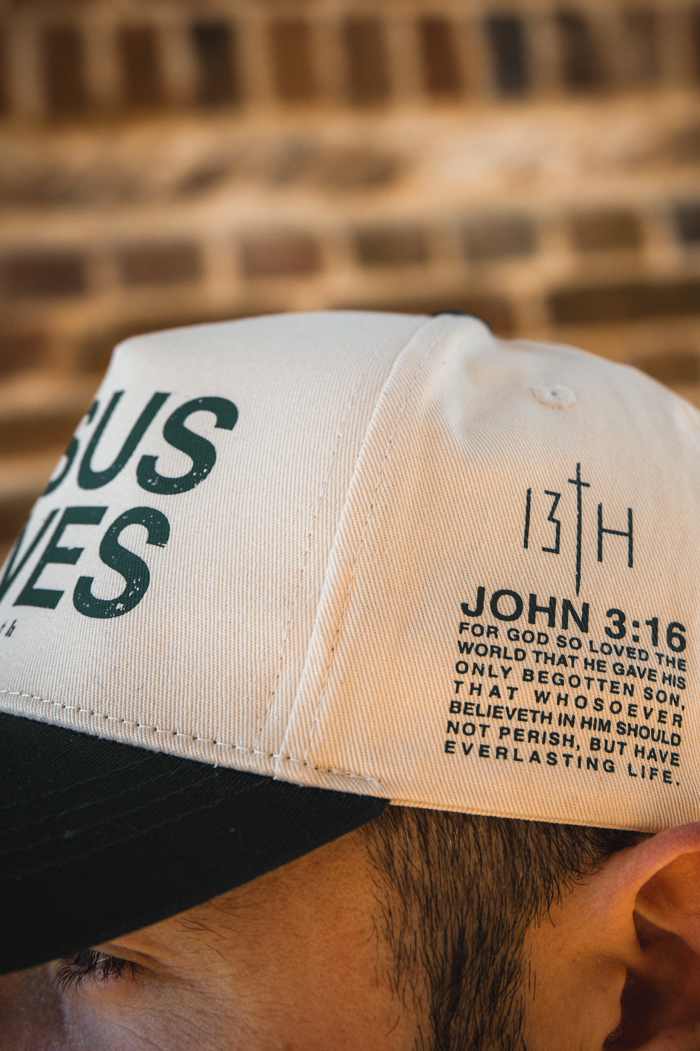 A close-up of the 13th Jesus Saves Ivory Snapback, featuring John 3:16 and the number 13 on the side. Part of a person’s face and ear are visible against a blurred brick background.