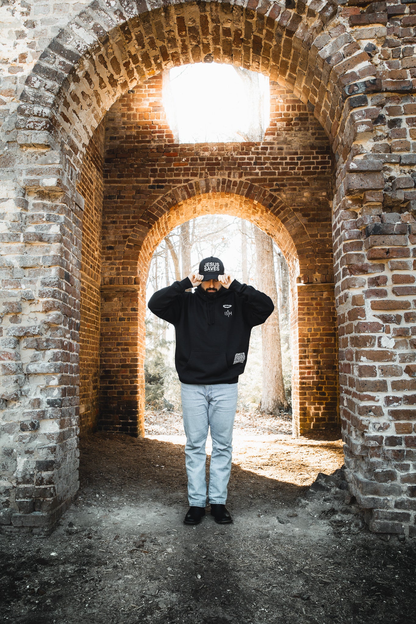 Wearing the 13th Jesus Saves Hoodie with light jeans, a person stands under a brick archway and adjusts their cap as sunlight pours in from above, casting a dramatic glow.