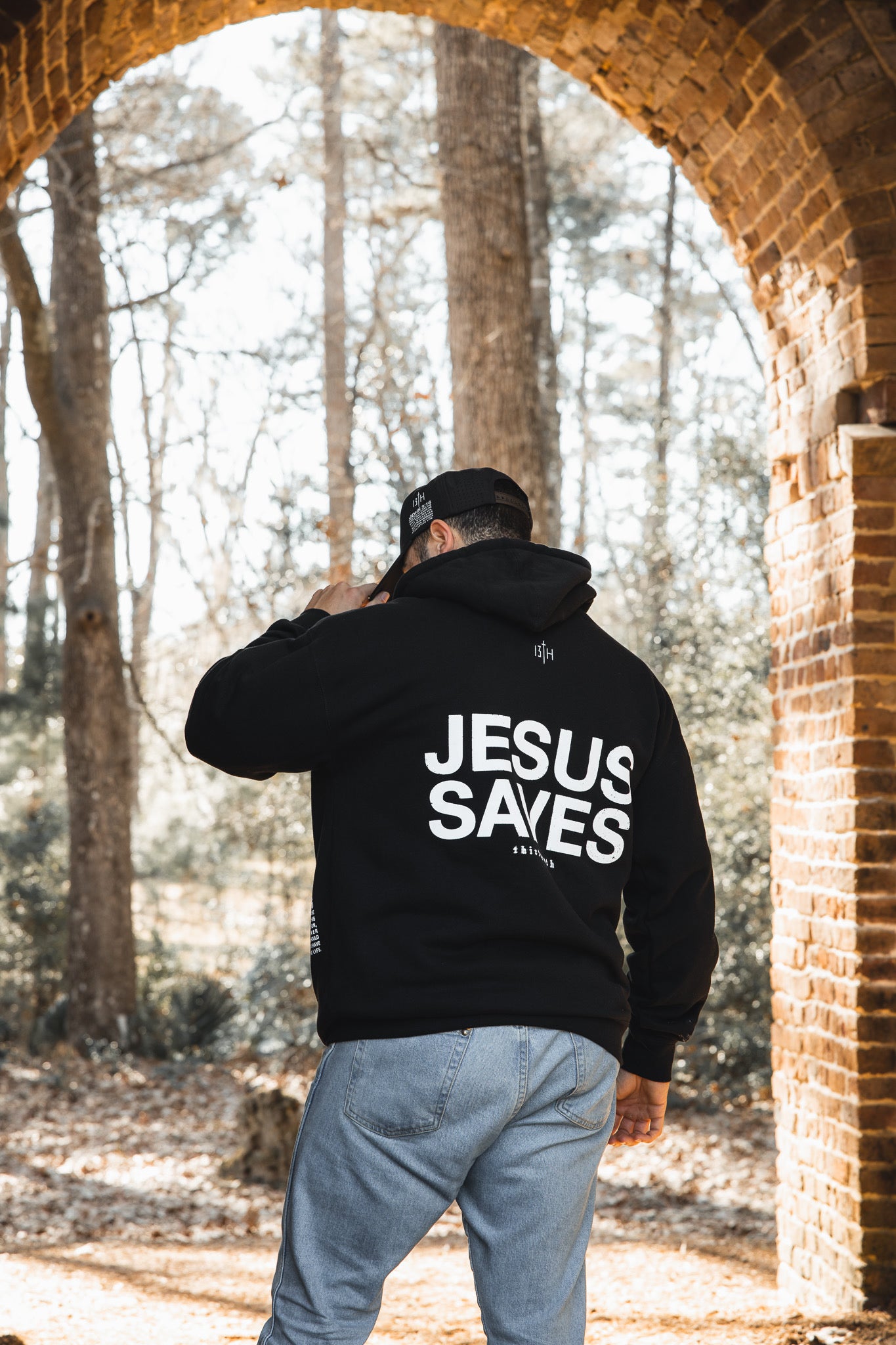 A person faces away, wearing the 13th Jesus Saves Hoodie in black with bold white lettering under a stone arch in the woods—an inspiring piece of faith apparel.
