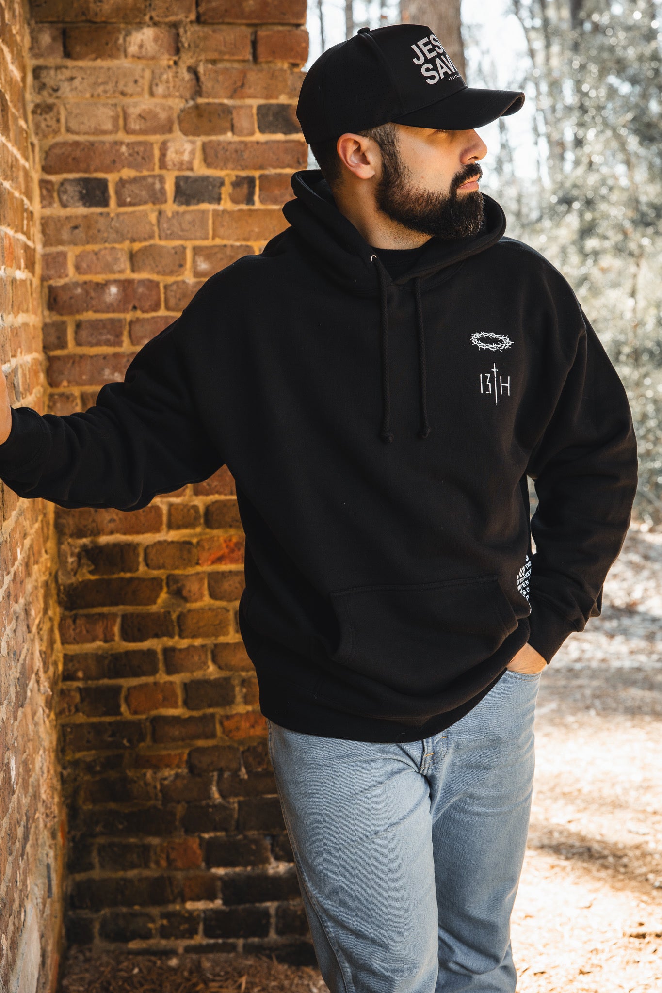 A bearded man in a black 13th Jesus Saves Hoodie and cap leans against a brick wall outdoors, one hand in his jeans pocket as sunlight filters through the trees behind him.