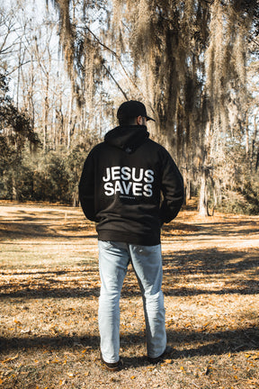 A person stands outdoors on fallen leaves, facing away, wearing the 13th Jesus Saves Hoodie and light blue jeans. Trees with hanging moss in the background emphasize this bold faith apparel in a peaceful natural setting.