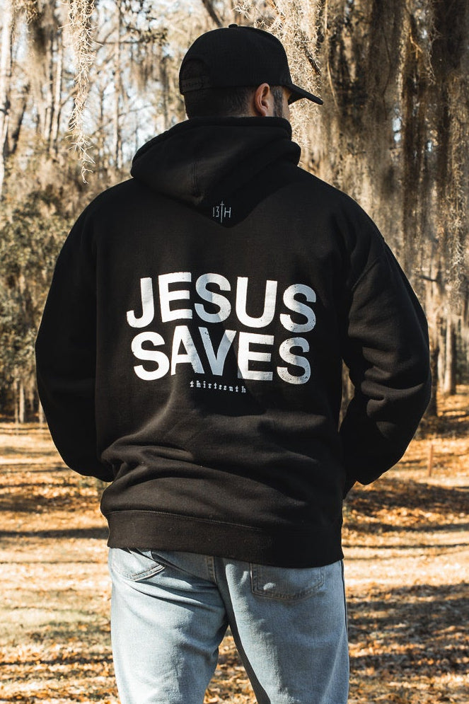 A person wearing the 13th Jesus Saves Hoodie, featuring bold “JESUS SAVES” text, stands outdoors amid trees and fallen leaves, facing away—showcasing this striking Christian faith apparel.