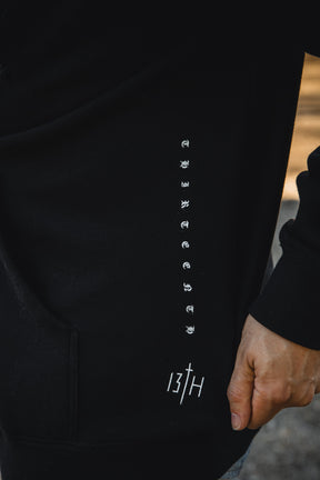 A person wears the Christ is King Premium Hoodie by 13th, a black hoodie featuring "THIRTEENTH" in white text down the front and "13TH" near the hem—a stylish, modern expression of faith.