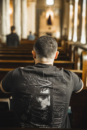 A man with short hair sits alone in a church pew, wearing the 13th He is Risen Premium Tee—a black shirt featuring Jesus crowned with thorns and "HE IS RISEN," boldly expressing his faith through this stylish Christian apparel.