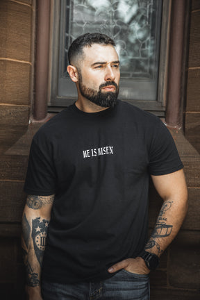 A bearded, tattooed man stands in front of a stone building wearing the 13th "He is Risen Premium Tee," looking to the side with a serious expression.