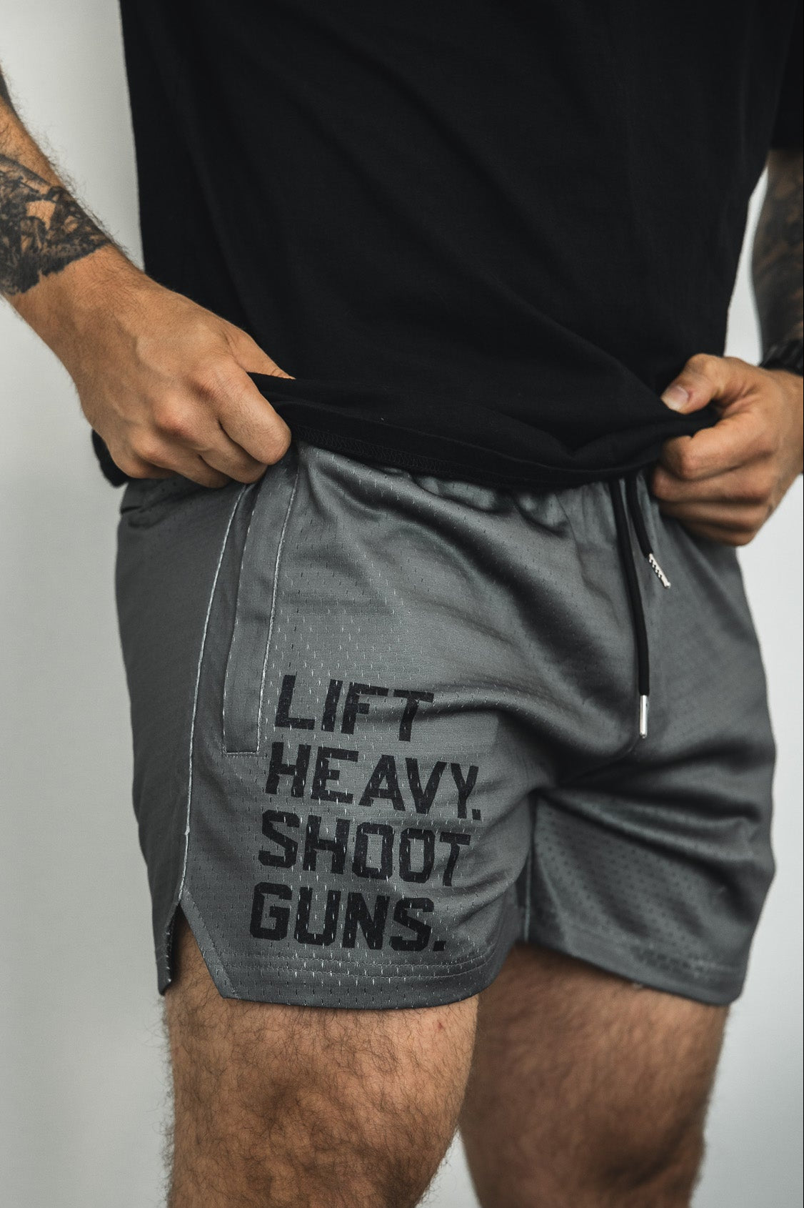 A person wears Tango Charlie Apparel Men's Mesh Lifestyle Shorts, 5.5" inseam, in OD Green with "LIFT HEAVY, SHOOT GUNS." on the left leg; their tattooed arms and hands rest near the waistband.