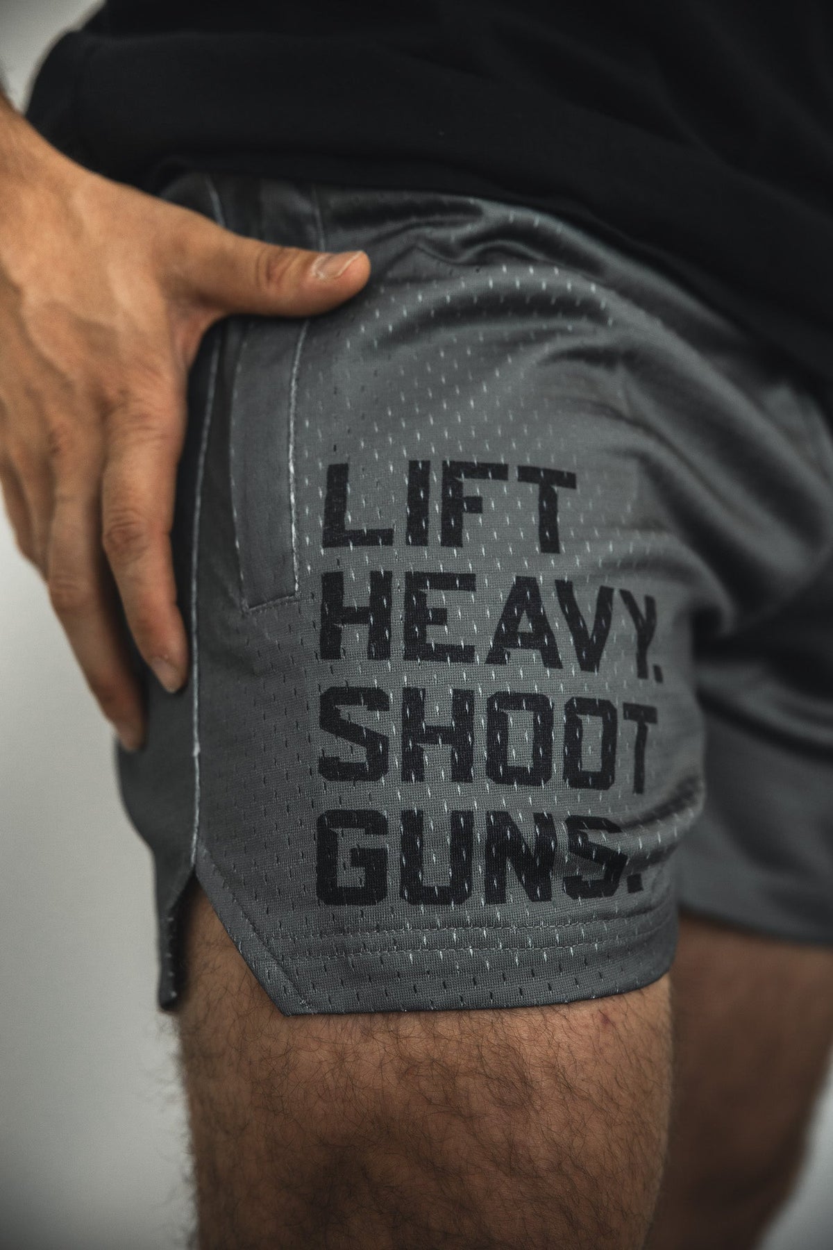 A close-up of a person wearing Tango Charlie Apparel Men's Mesh Lifestyle Shorts | 5.5"- LHSG OD Green, with "LIFT HEAVY. SHOOT GUNS." in bold black letters on the side and a hand resting near the pocket, showcasing these shorts.