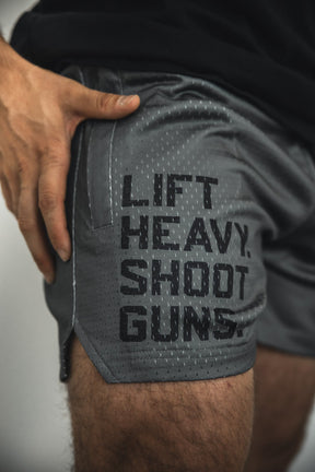 A close-up of a person wearing Tango Charlie Apparel Men's Mesh Lifestyle Shorts | 5.5"- LHSG OD Green, with "LIFT HEAVY. SHOOT GUNS." in bold black letters on the side and a hand resting near the pocket, showcasing these shorts.