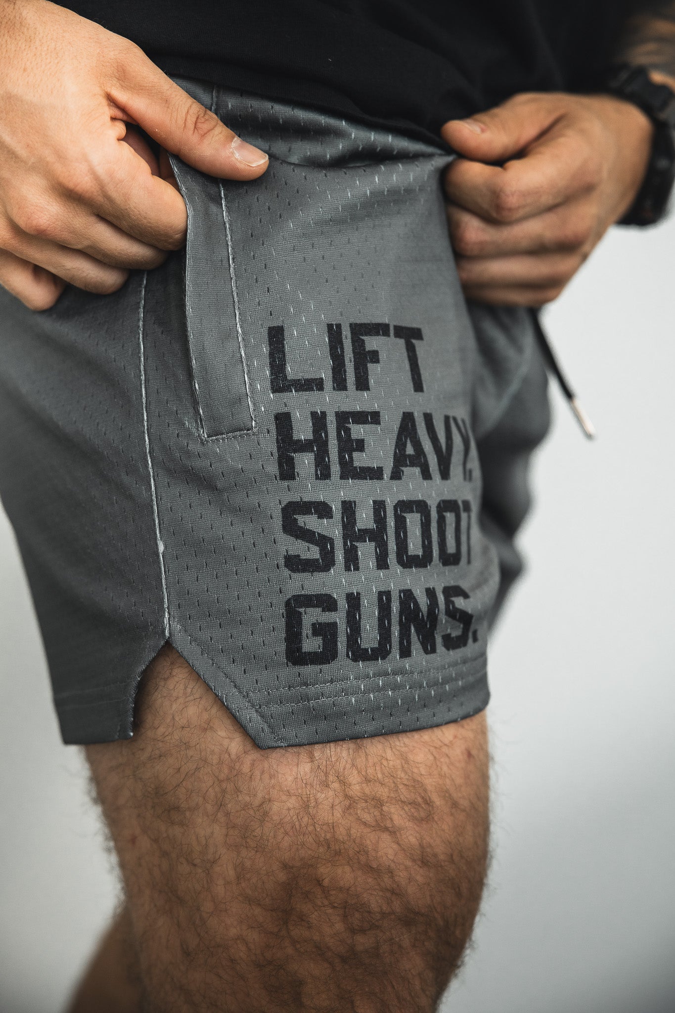 A person wears Tango Charlie Apparel Men's Mesh Lifestyle Shorts | 5.5"- LHSG OD Green, featuring LIFT HEAVY. SHOOT GUNS. printed in black on the side, holding the waistband with both hands.