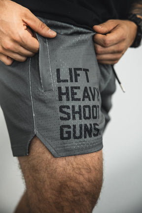 A person wears Tango Charlie Apparel Men's Mesh Lifestyle Shorts | 5.5"- LHSG OD Green, featuring LIFT HEAVY. SHOOT GUNS. printed in black on the side, holding the waistband with both hands.