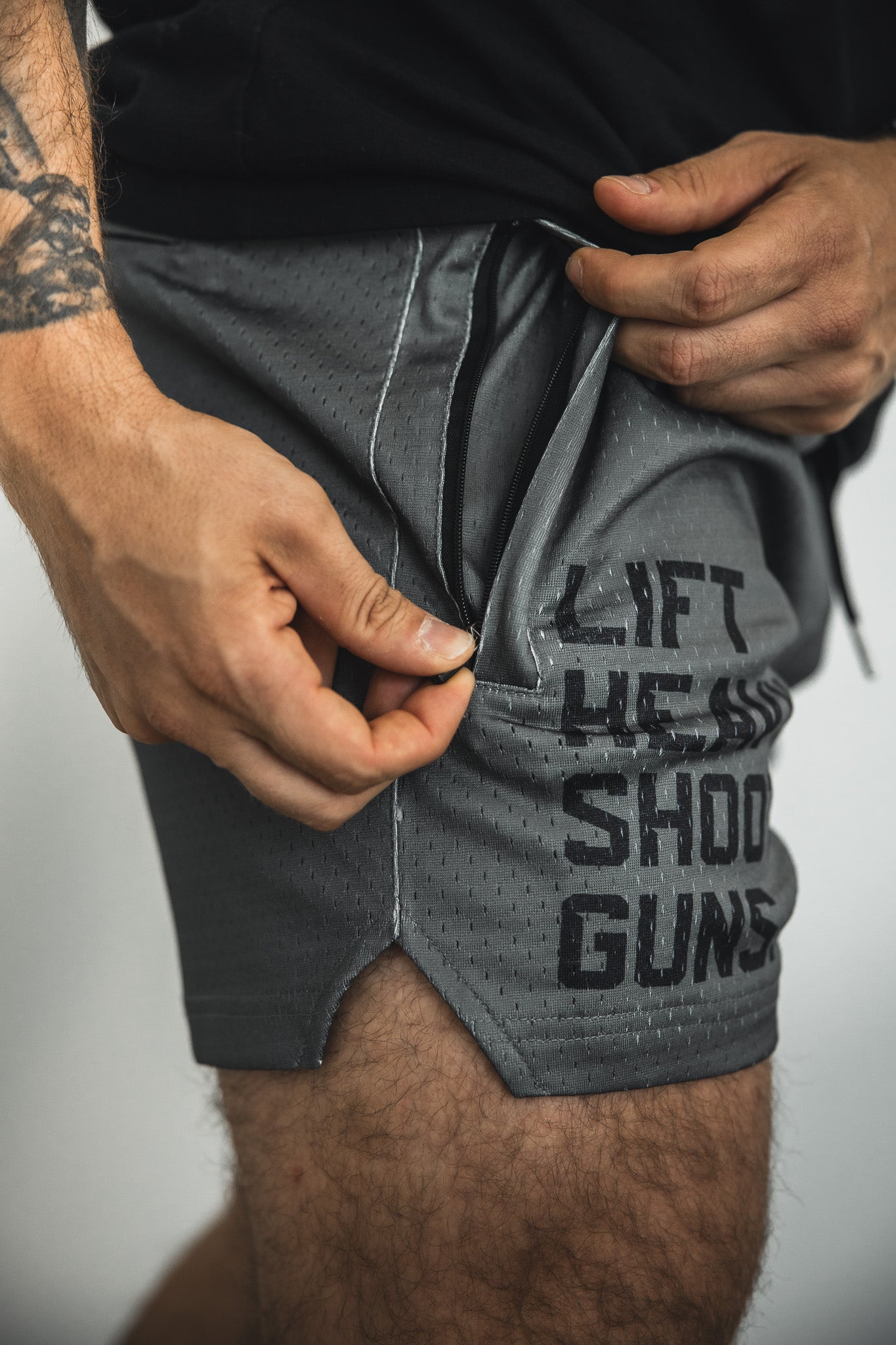 A person with a visible arm tattoo opens the small zippered pocket on Tango Charlie Apparel’s Men's Mesh Lifestyle Shorts | 5.5" in LHSG OD Green, featuring “LIFT HE SHO GUNS” printed on the side.