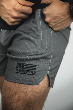 A close-up of a person wearing Tango Charlie Apparel Men's Mesh Lifestyle Shorts in OD Green, featuring perforated fabric, a 5.5” inseam, black flag logo, and "BORN UNITED" on the left leg as they adjust the waistband.