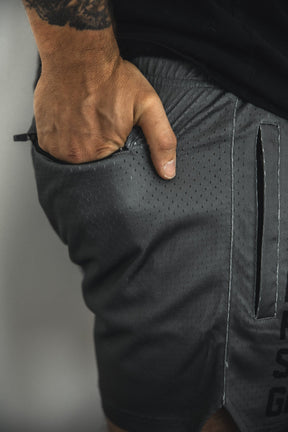 A close-up of a person wearing Tango Charlie Apparel Men's Mesh Lifestyle Shorts | 5.5"- LHSG OD Green, featuring perforated detailing, visible stitching, a hand in the pocket, and a forearm tattoo.