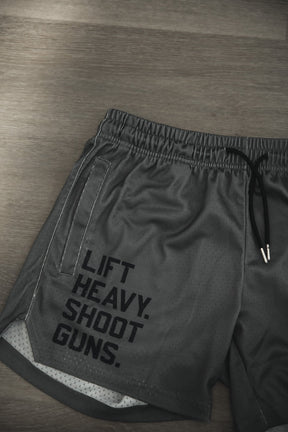 Tango Charlie Apparel Men's Mesh Lifestyle Shorts | 5.5"- LHSG OD Green feature black drawstrings and bold "LIFT HEAVY. SHOOT GUNS." text on the left leg, shown laid flat on a wooden surface.