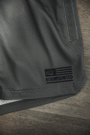 Close-up of Tango Charlie Apparel Men's Mesh Lifestyle Shorts (5.5"- LHSG OD Green) showing a “BORN UNITED” patch with a flag design. These athletic shorts offer everyday comfort and style on a wooden surface.