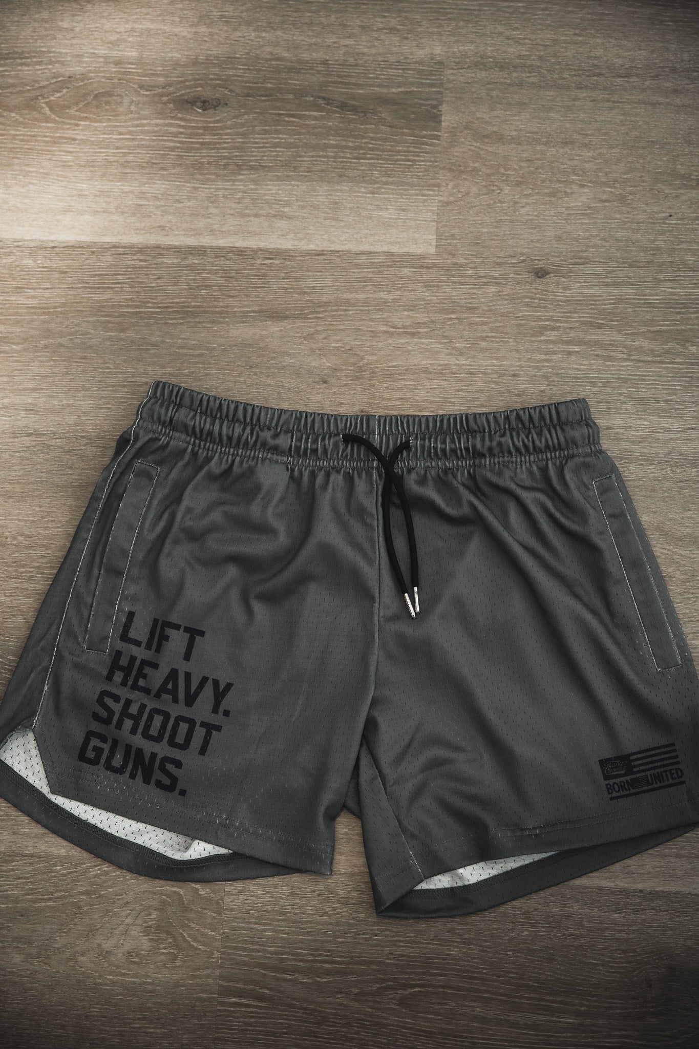 The Tango Charlie Apparel Men's Mesh Lifestyle Shorts | 5.5"- LHSG OD Green feature bold "LIFT HEAVY. SHOOT GUNS." text on the left leg, black drawstrings, and are perfect for workouts or everyday wear.