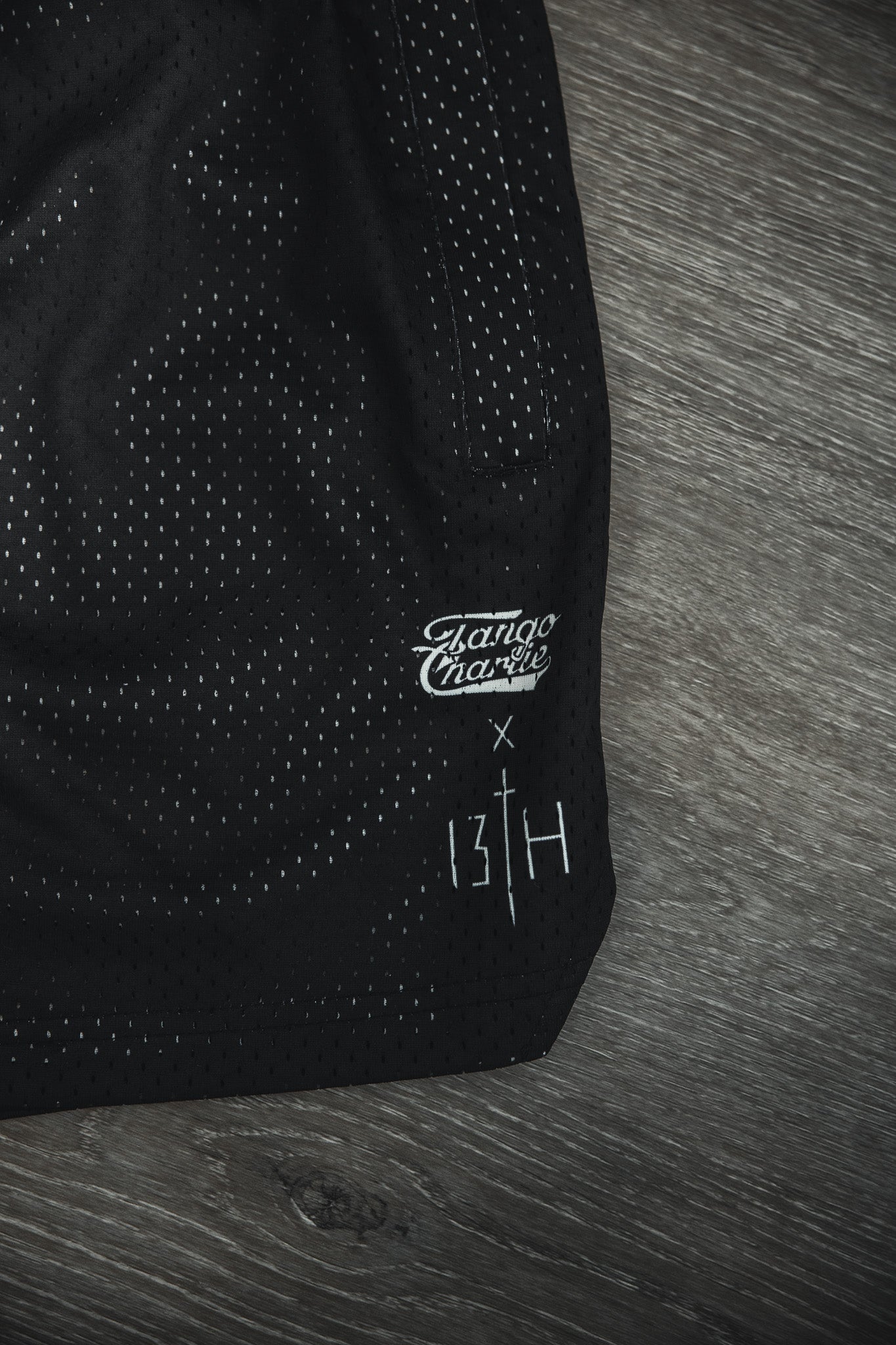 Close-up of Tango Charlie Apparel Men's Mesh Lifestyle Shorts | 5.5"- Lift Heavy Pray Hard Black, shown on wood, featuring collaboration details and a stitched white cross—ideal for gym or everyday wear.
