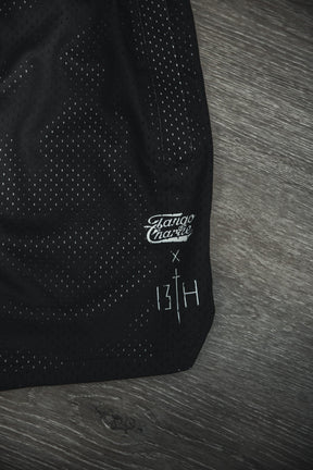 Close-up of Tango Charlie Apparel Men's Mesh Lifestyle Shorts | 5.5"- Lift Heavy Pray Hard Black, shown on wood, featuring collaboration details and a stitched white cross—ideal for gym or everyday wear.
