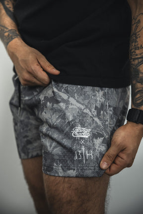 A person wearing a black shirt and Tango Charlie Apparel Men's Mesh Lifestyle Shorts | 5.5"- Lift Heavy Pray Hard Deadwood, featuring gray camo, white printed logos and text, with tattoos on their arms and a black watch.