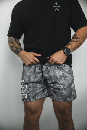 A person wearing a black t-shirt and Tango Charlie Apparel Men's Mesh Lifestyle Shorts | 5.5"- Lift Heavy Pray Hard Deadwood stands against a plain background, holding the waistband, with tattoos on both arms and a wristwatch.