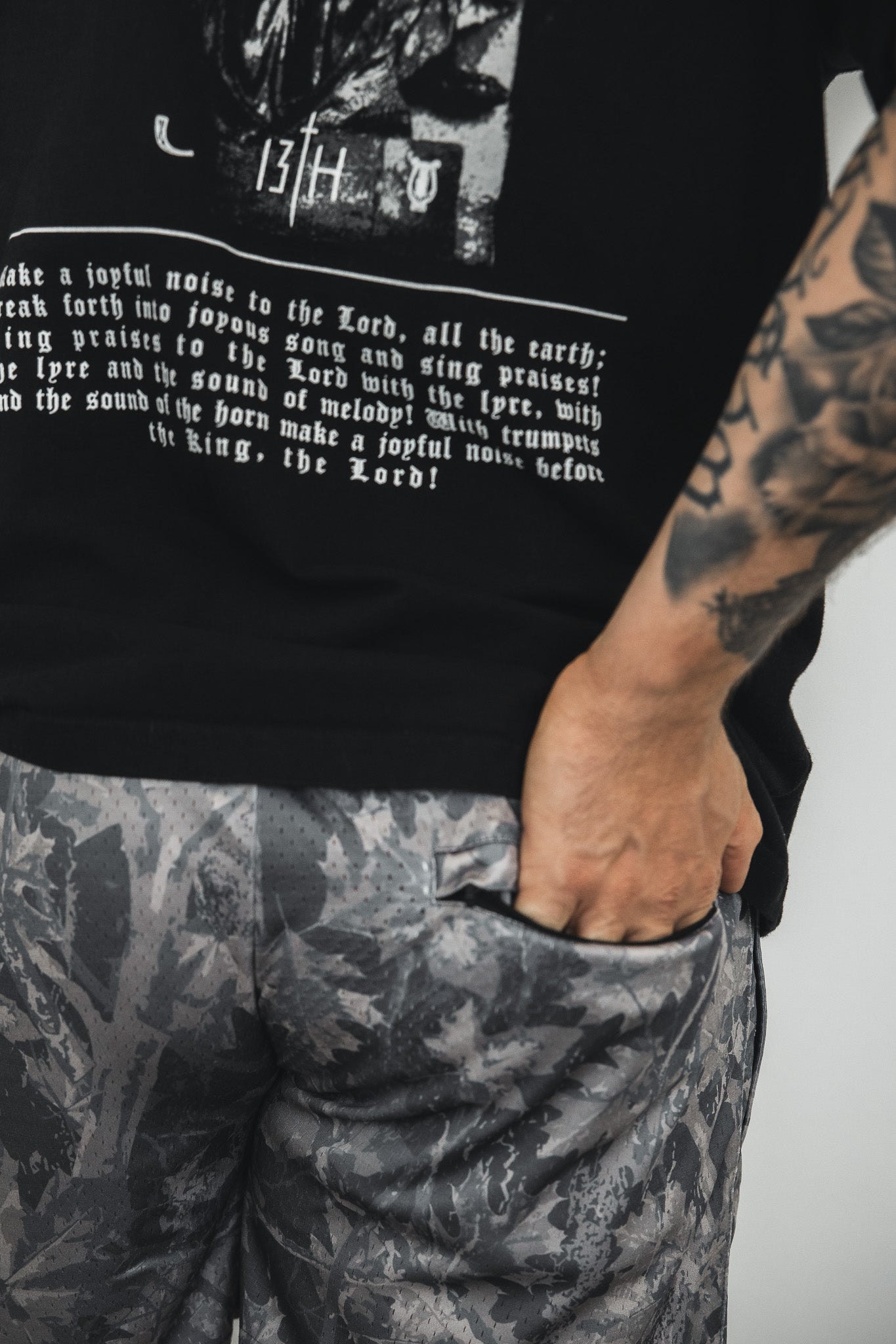 A person with a tattooed arm stands confidently with one hand in their pocket, wearing a black shirt with a religious message and Tango Charlie Apparel Men's Mesh Lifestyle Shorts | 5.5"- Lift Heavy Pray Hard Deadwood.