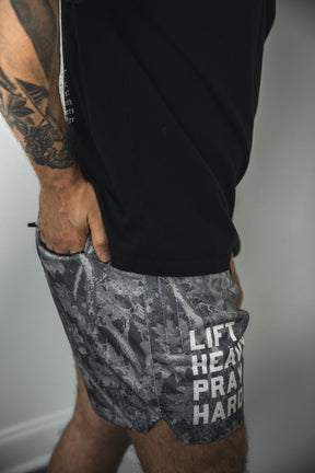 A tattooed man in a black shirt and Tango Charlie Apparel Men's Mesh Lifestyle Shorts | 5.5" - Lift Heavy Pray Hard Deadwood, featuring "LIFT HEAVY PRAY HARD" on the side, stands with one hand in his pocket.