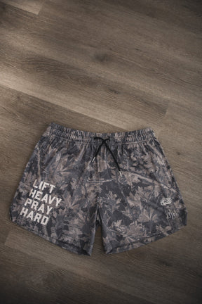 The Tango Charlie Apparel Men's Mesh Lifestyle Shorts | 5.5"- Lift Heavy Pray Hard Deadwood feature a camo pattern and "LIFT HEAVY PRAY HARD" in white on the left leg, shown laid flat on a wooden floor.