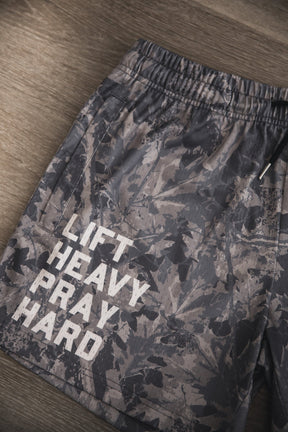 A pair of Tango Charlie Apparel Men's Mesh Lifestyle Shorts | 5.5"- Lift Heavy Pray Hard Deadwood with a camouflage pattern and "LIFT HEAVY PRAY HARD" in bold white letters is displayed on a wooden surface.