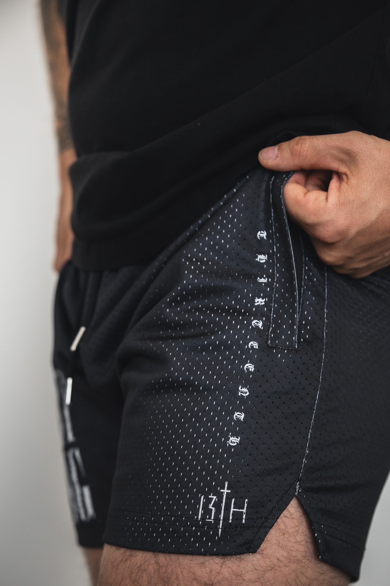 A person wears 13th Men's Mesh Lifestyle Shorts | 5.5"–Christ is King, featuring black mesh fabric with white stitching and text on the side and hem, holding the waistband; their upper body in a black shirt is partially visible.