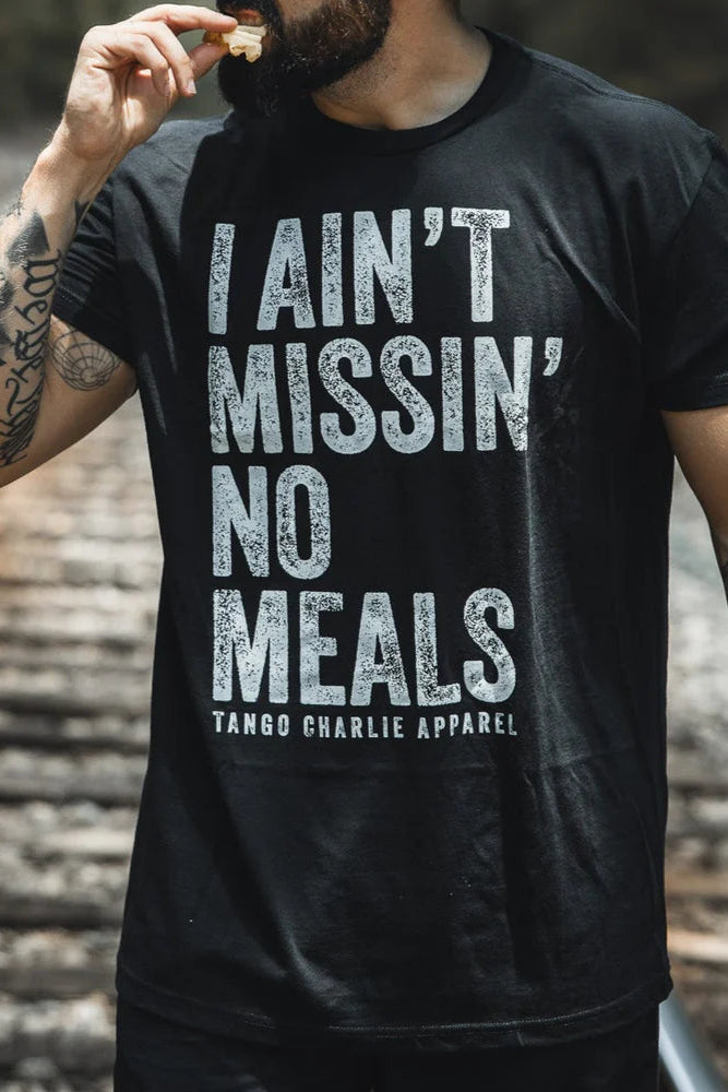 Sporting the I Ain’t Missin’ No Meals Tee by Tango Charlie Apparel, someone enjoys a snack outdoors—showing they’re always hungry.