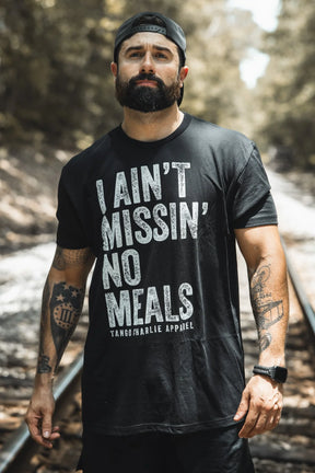 A bearded man in a backward cap wears the black "I Ain’t Missin’ No Meals Tee" by Tango Charlie Apparel—ideal for the always hungry—as he stands on railroad tracks with blurred trees behind him.