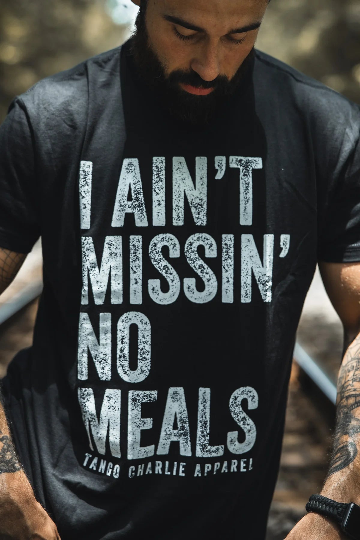 A bearded, tattooed man wears the I Ain’t Missin’ No Meals Tee by Tango Charlie Apparel. This comfy black cotton-poly blend shirt boasts bold white lettering—ideal for anyone who’s always hungry.