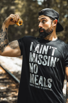 A bearded man in a backward cap and the "I Ain’t Missin’ No Meals Tee" by Tango Charlie Apparel holds up food outdoors, eyeing it with surprise—proving he’s always hungry and ready for more.