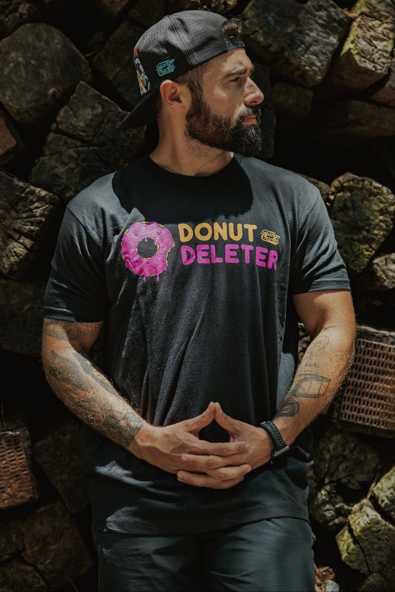 A bearded man wears the Tango Charlie Apparel Donut Deleter Tee—made from a comfy cotton-poly blend—standing before stacked logs with tattooed arms visible and a backwards cap for style.