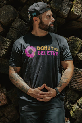 A bearded man wears the Tango Charlie Apparel Donut Deleter Tee—made from a comfy cotton-poly blend—standing before stacked logs with tattooed arms visible and a backwards cap for style.