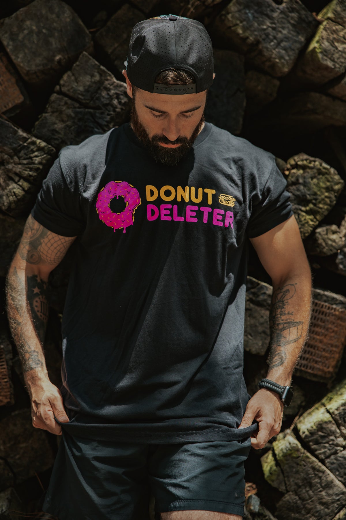 A man in a black backwards cap and shorts looks down at his Donut Deleter Tee by Tango Charlie Apparel, a black cotton-poly shirt featuring "DONUT DELETER" with a pink frosted donut as the O. Stacked firewood fills the background.