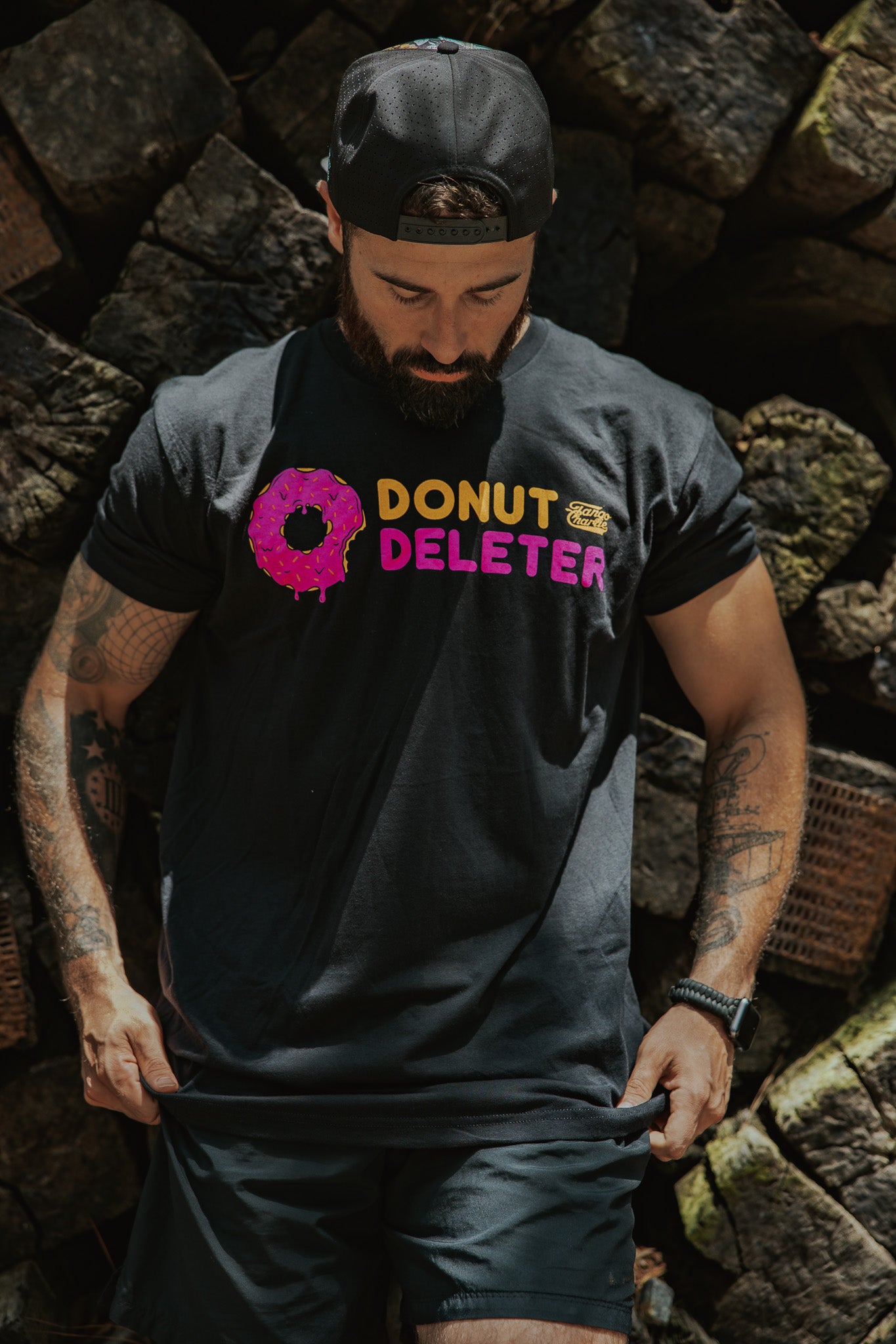 A man in a black backwards cap and shorts looks down at his Donut Deleter Tee by Tango Charlie Apparel, a black cotton-poly shirt featuring "DONUT DELETER" with a pink frosted donut as the O. Stacked firewood fills the background.