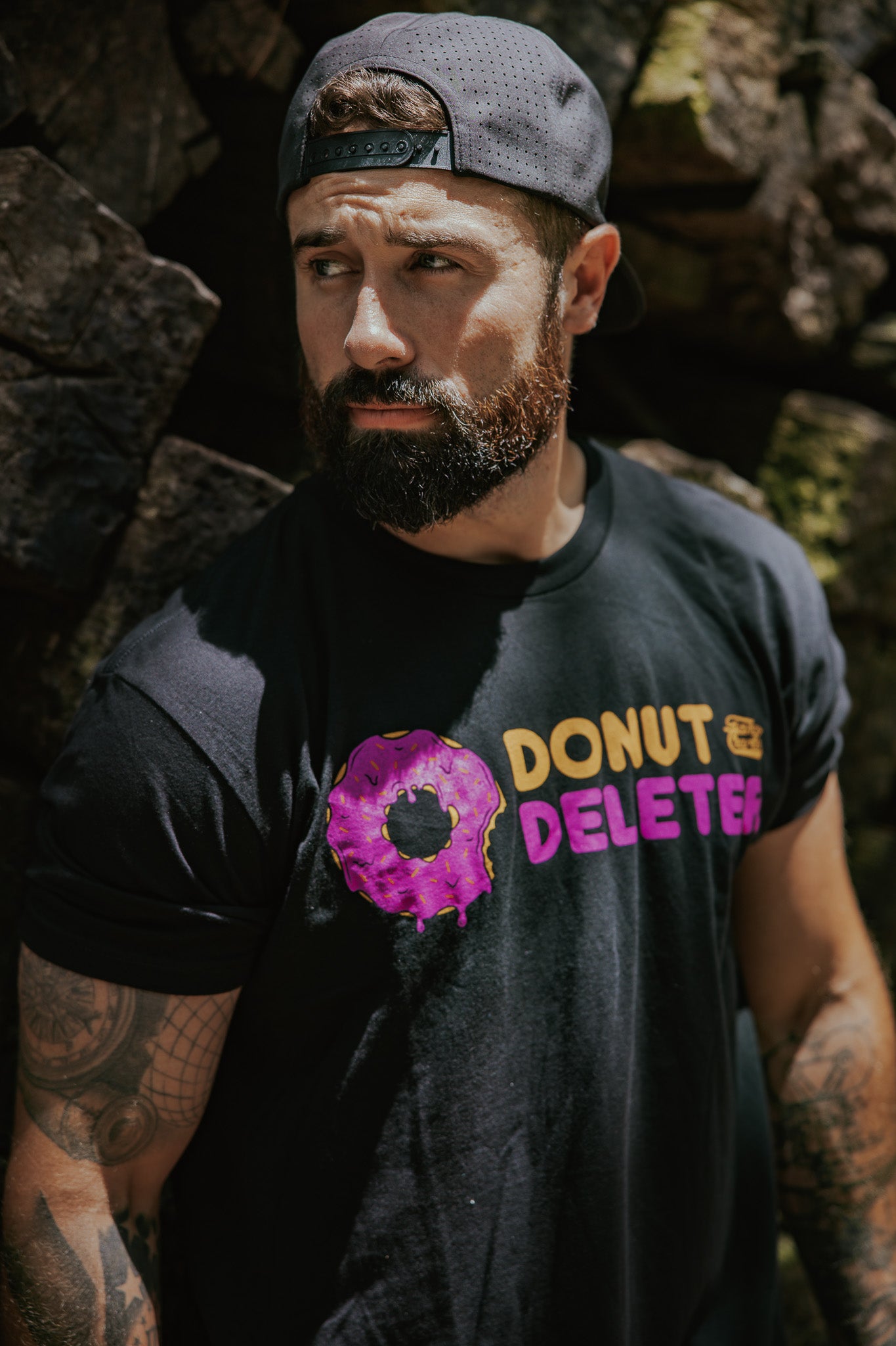 A bearded man in a backwards cap stands by rocks outdoors, wearing the Tango Charlie Apparel Donut Deleter Tee—a black cotton-poly shirt with a purple donut and “DONUT DELETER” in yellow and pink letters.