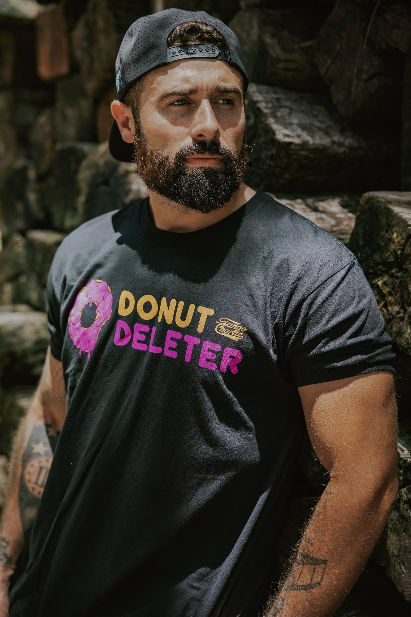 A bearded man in a black backward cap and Donut Deleter Tee by Tango Charlie Apparel stands against a stone wall, looking to the side with a serious expression, his laid-back yet focused vibe highlighted by the soft cotton poly blend shirt.