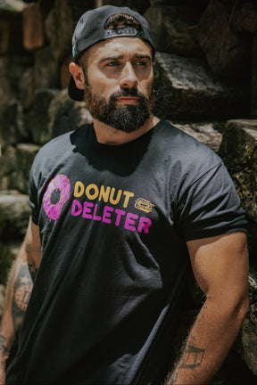 A bearded man in a black backward cap and Donut Deleter Tee by Tango Charlie Apparel stands against a stone wall, looking to the side with a serious expression, his laid-back yet focused vibe highlighted by the soft cotton poly blend shirt.